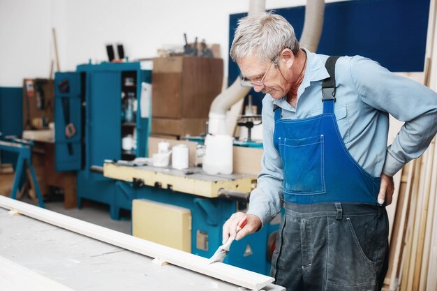 An elderly cabinetmaker in overalls and glasses paints a wooden\
board with a brush on a workbench in a carpentry shop