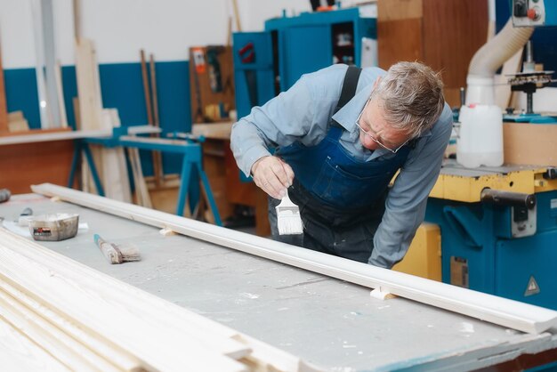 An elderly cabinetmaker in overalls and glasses paints a wooden board with a brush on a workbench in a carpentry shop