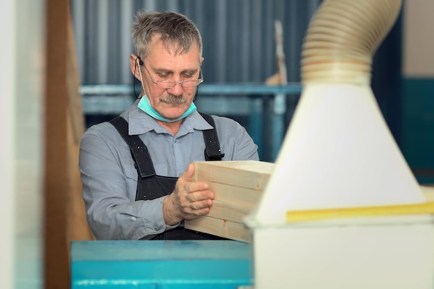 An elderly carpenter of caucasian appearance works in a\
carpentry workshop processing of boards on a lathe