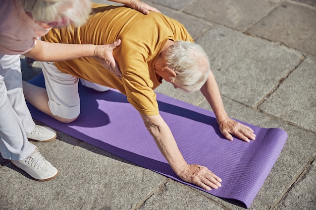 Elderly Caucasian beginner yogi performing a back stretch exercise assisted by a fit gray-haired lady