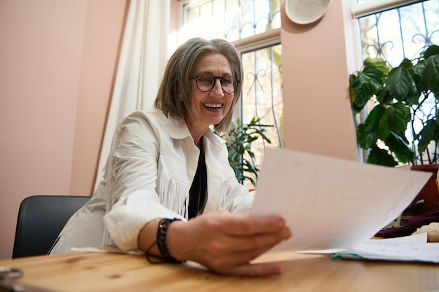 An elderly Caucasian grayhaired woman couturier and fashion designer looks at sketches for clothes sitting at a wooden desk in her creative office