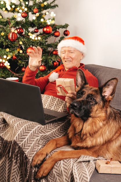 Elderly caucasian man in Santa hat with laptop sitting on a sofa near a Christmas tree with gift box and German Shepherd dog chatting with relatives online. Self-isolation, holiday mood.