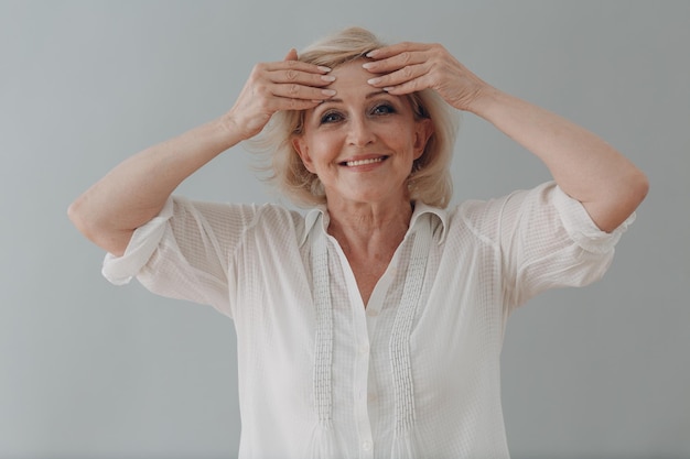 Elderly caucasian old aged woman gray haired portrait doing facebuilding yoga face gymnastics yoga self massage