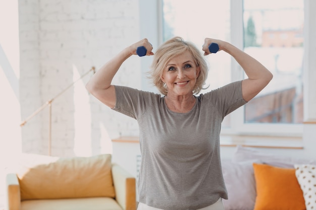 Elderly caucasian old aged woman portrait gray haired showing biceps muscles doing exercises with dumbbells in casual wear at home apartment living room