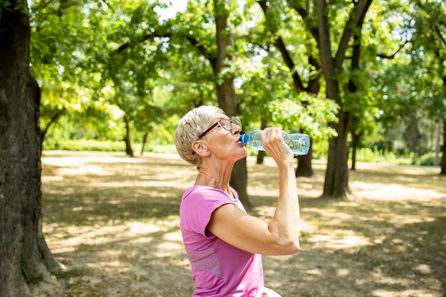 Donna caucasica anziana che si prende una pausa e beve acqua dopo un lungo allenamento in natura.