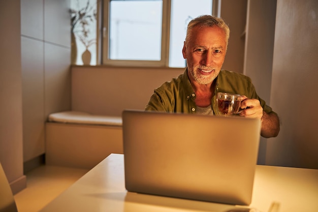 Elderly citizen smiling while having tea near his laptop