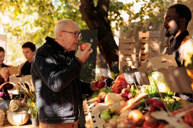Пожилой клиент нюхает био-яблоки и стоит возле прилавка фермерского продовольственного рынка с разнообразными свежими фруктами и овощами, не содержащими пестицидов. Старик покупает органические продукты местного производства.