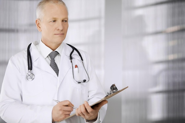 Elderly confident doctor standing straight and using clipboard in clinic Perfect medical service Medicine and healthcare concept