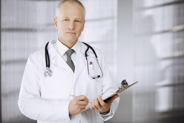 Elderly confident doctor standing straight and using clipboard in clinic. Perfect medical service. Medicine and healthcare concept.