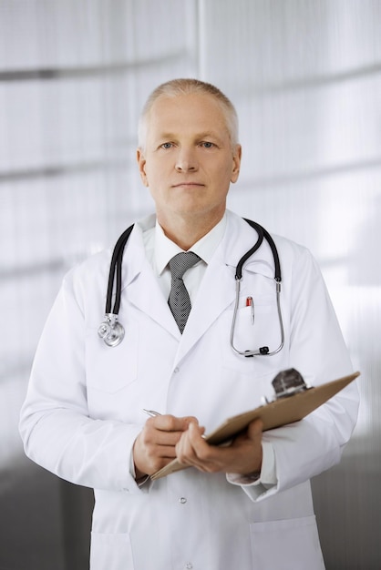 Elderly confident doctor standing straight and using clipboard in clinic. Perfect medical service. Medicine and healthcare concept.