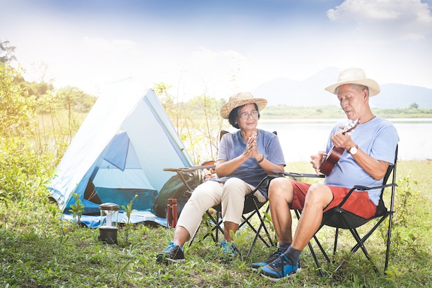 elderly couple camping in a forest by the river. Both sit on a chair and play music. Happy life in retirement. Senior community concepts