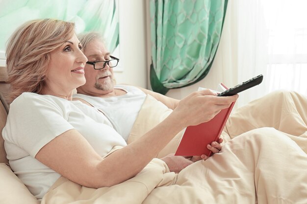 Elderly couple reading book and watching TV while resting in bed at home