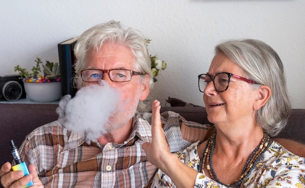 Elderly couple sitting indoors, the husband smokes an electronic cigarette and the wife is annoyed by the smoke