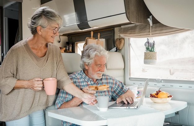 Elderly couple in travel vacation inside a camper van enjoying\
breakfast together happy caucasian senior couple in van life while\
browsing on laptop