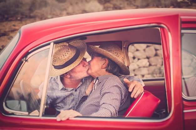 Foto coppia di anziani con cappello, con gli occhiali, con i capelli grigi e bianchi, con camicia casual, su un'auto rossa d'epoca in vacanza godendosi il tempo e la vita con un allegro cellulare sorridente immerso nella ventosa sp