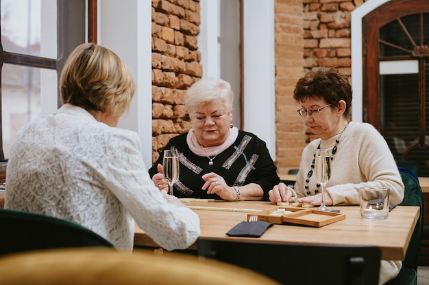 Пожилые темноволосые и светловолосые женщины, одетые в черно-белые куртки, сидят в уютном кафе за столом со свечой, разговаривают и празднуют встречу с друзьями