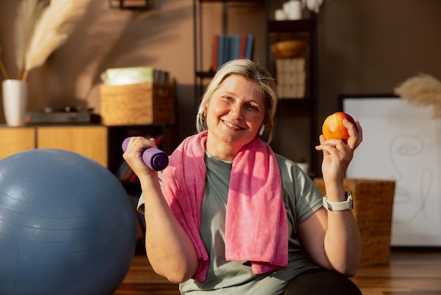 Elderly delighted mother training alone at home sitting on\
floor holding dumbbells