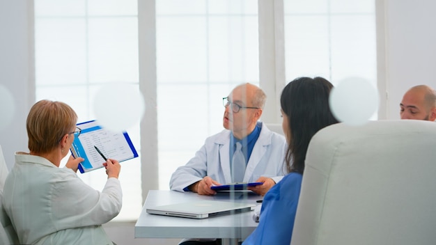 Elderly doctor having medical conference in hospital meeting room discussing medical problems pointing on clipboard presenting list of sick patients. Team of doctors presenting symptoms of disease