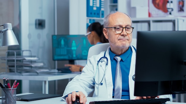 Elderly doctor working on PC in modern private clinic with glass walls. Hospital professionals in health industry staff at work in medicine medical practitioner specialist in unifor at desk job