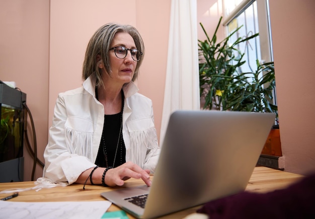 Elderly European woman fashion designer working in clothing design and tailoring studio and using laptop while designing new garment