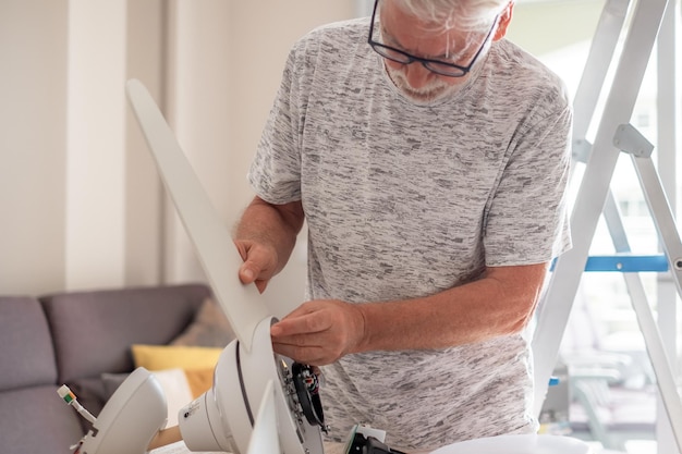 Elderly experienced electrician man works to install a paddle fan on the ceiling at home