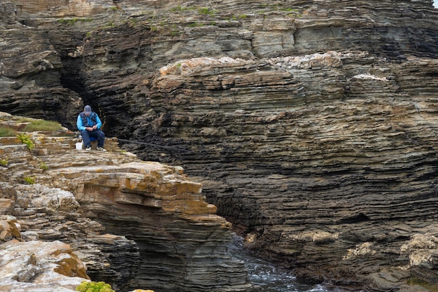 年配の漁師が崖の岩の上に座ってフックとロッドを準備している