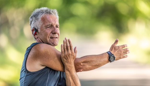 Elderly fit man stretches his arm on a summer day outdoors, wearing watches and earphones.