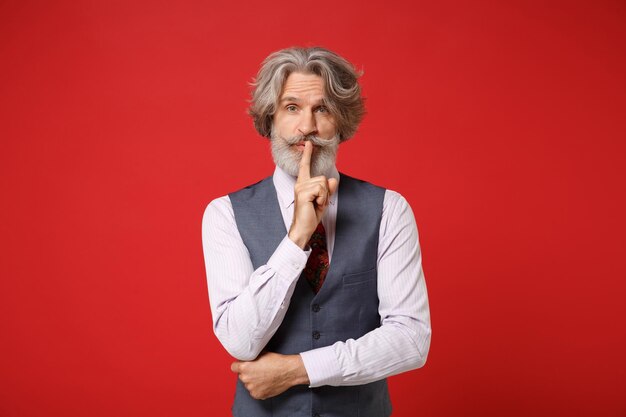 Elderly gray-haired mustache bearded man in classic shirt vest\
tie posing isolated on red background. people lifestyle concept.\
mock up copy space. say hush be quiet with finger on lips shhh\
gesture.
