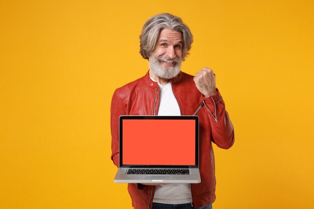 Elderly gray-haired mustache bearded man in red leather jacket isolated on yellow background. People lifestyle concept. Mock up copy space. Hold laptop computer with empty screen doing winner gesture.