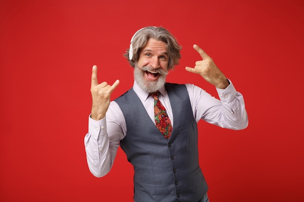 Elderly gray-haired mustache bearded man in shirt vest tie\
isolated on red wall background. people lifestyle concept. mock up\
copy space. listen music with headphones, depicting heavy metal\
rock sign.