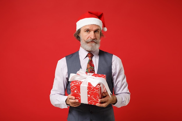 Elderly gray-haired mustache bearded santa man in christmas hat\
shirt vest tie isolated on red background. new year 2020\
celebration concept. mock up copy space. hold present box with gift\
ribbon bow.