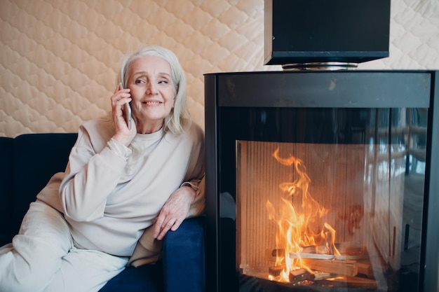 Elderly gray haired woman sitting on sofa and talking smartphone in living room with fireplace