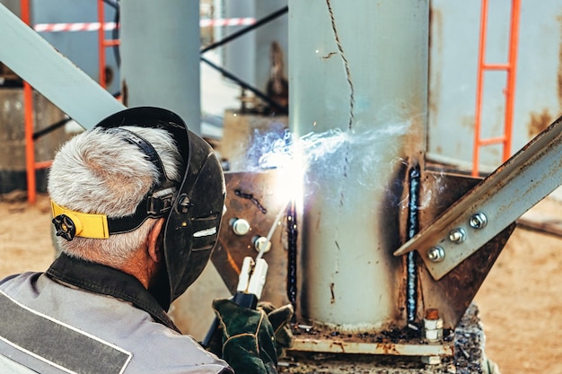 An elderly grayhaired grayhaired welder assembles metal\
structures at a construction site arc welding work sparks from\
welding closeup