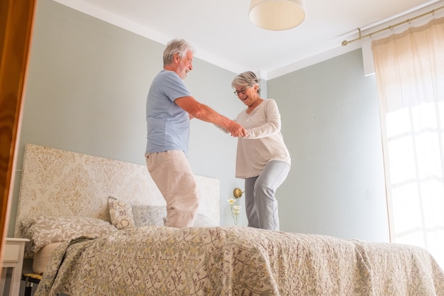 Elderly happy senior couple holding hands and dancing together on bed at home. Carefree active senior heterosexual couple holding hands and dancing together on bed at home