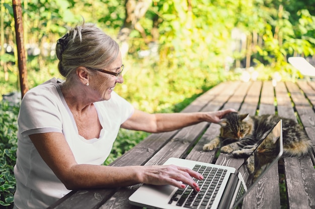 庭で屋外のラップトップコンピューターでオンラインで作業している家猫と年配の幸せな年配の女性。リモートワーク