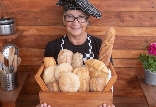 Elderly happy woman holding a wooden basket with a large selection of fresh bread made with different flours Background in recycled wood