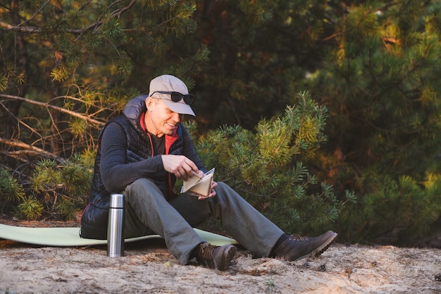 Elderly hiker eating a sandwich and drinking a hot drink sitting on a tourist rug in forest