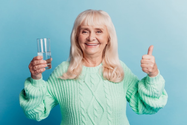 Пожилая женщина держит стакан чистой минеральной воды, показывает жест на синем фоне
