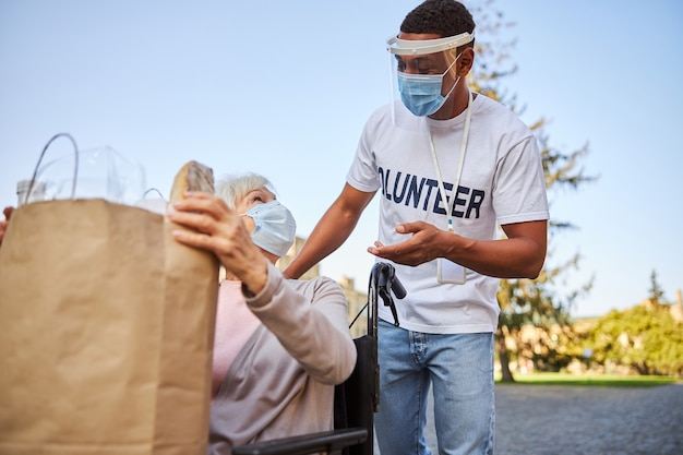 Photo elderly lady in medical mask turning beck in her wheelchair and looking at the young man in a protective shield