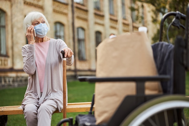 Elderly lady with a walking stick sitting outdoors and looking into the distance while talking on the phone