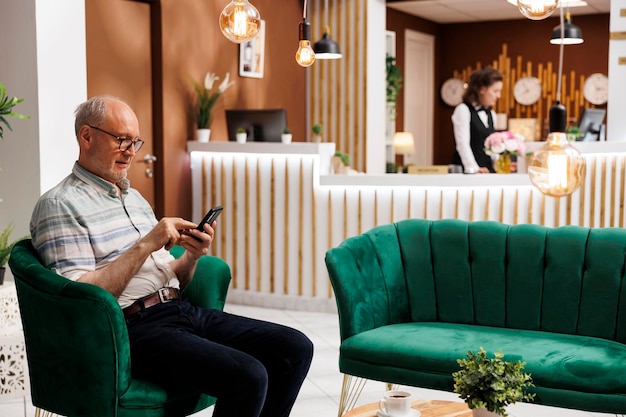 Elderly male customer seated on sofa using mobile device while waiting for checkin in hotel lobby Retired senior man surfing the net for holiday activities on his smartphone in lounge area