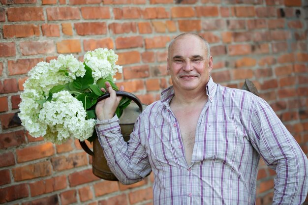 Elderly male florist A man holds a watering can with a bouquet of flowers against a brick wall