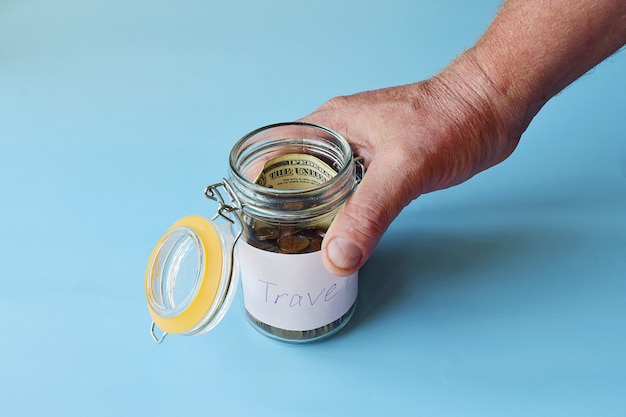 Elderly male hand holds a glass jar with money savings,
banknotes and coins on a blue background in sunlight. travel money
savings concept