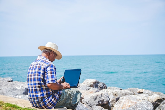 海沿いの職場で座っているラップトップコンピューターを運ぶ老人