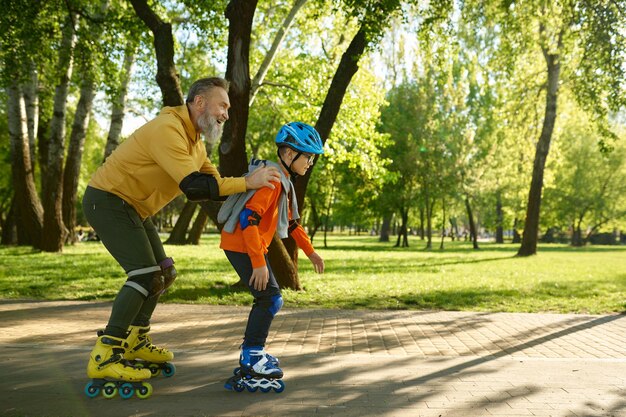 Пожилой мужчина занимается спортом в солнечном летнем парке со своим маленьким сыном. Счастливый обрадованный отец следует за мальчиком, катающимся на роликовых коньках