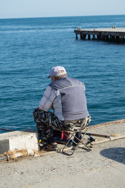 An elderly man fisherman sitting on a parapet by the sea and fishing on a fishing rod