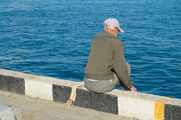 Un uomo anziano pescatore seduto su un parapetto in riva al mare e la pesca su una canna da pesca