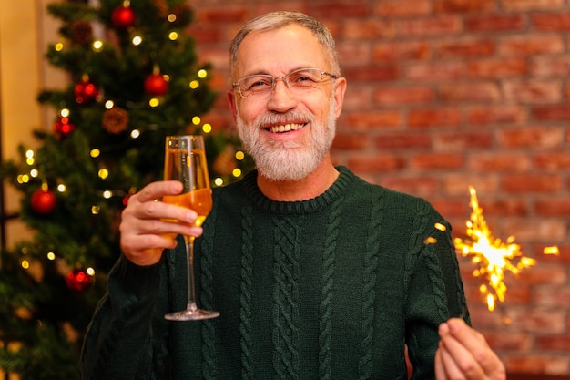 An elderly man in a green knitted sweater raising a toast with champagne near the Christmas tree