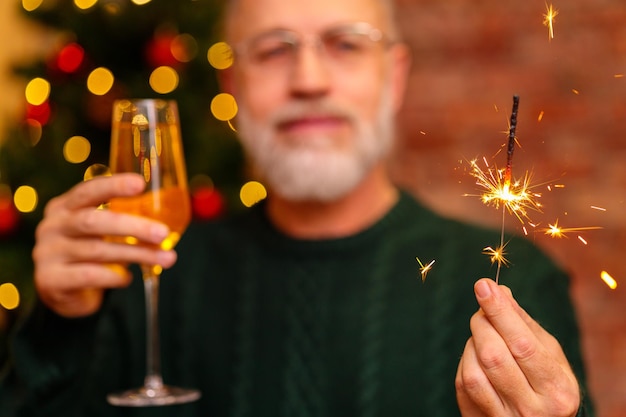 Un uomo anziano con un maglione lavorato a maglia verde che brinda con champagne vicino all'albero di natale