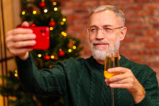 Un uomo anziano in una maglia verde che parla con la famiglia tramite la connessione video del telefono vicino all'albero di natale